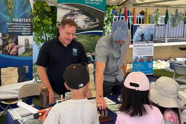 Two adults wearing clothes with the NOAA logo stand behind an outreach table