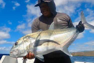 A smiling fisherman holds a nearly 35-inch-long silvery fish in his hands while on a boat in the ocean.