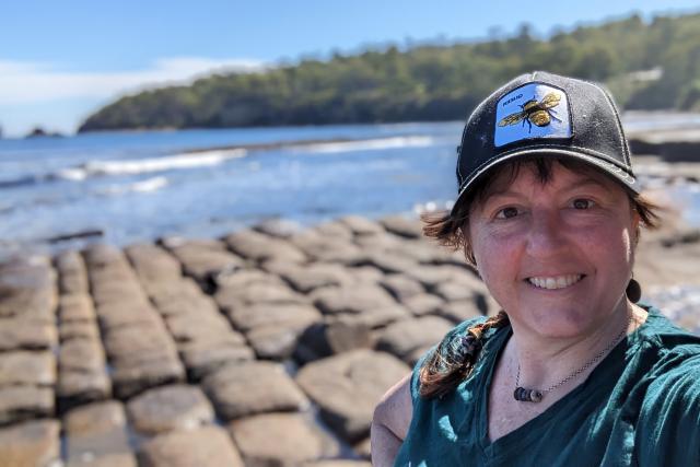 A woman wearing a black baseball cap with a bee on it poses in front of the ocean.