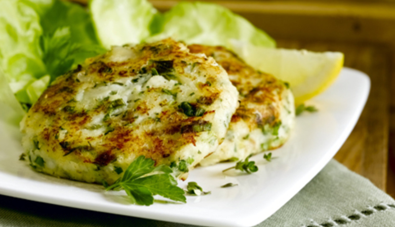 a side view of a square white plate sitting on a grey-green cloth and wooden table. the plate has two round cod-potato cakes in the foreground with a lemon wedge and lettuce in the background. the dish is garnished with leafy hearbs